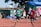 Several young athletes are sprinting on a track in what appears to be a race, with spectators and other participants visible in the background. The runners are wearing numbered bibs and colorful sports attire, competing intensely under clear skies.