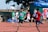 Several young athletes are sprinting on a track in what appears to be a race, with spectators and other participants visible in the background. The runners are wearing numbered bibs and colorful sports attire, competing intensely under clear skies.