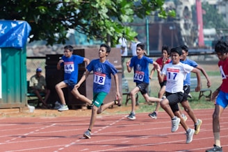 A group of young athletes are mid-race on a track, displaying focus and determination. They wear numbered bibs and sport attire with several in blue shirts. The background includes trees and observers.
