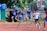 A group of young athletes are mid-race on a track, displaying focus and determination. They wear numbered bibs and sport attire with several in blue shirts. The background includes trees and observers.