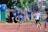 A group of young athletes are mid-race on a track, displaying focus and determination. They wear numbered bibs and sport attire with several in blue shirts. The background includes trees and observers.