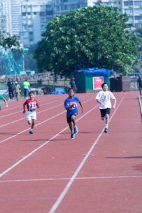 Several young boys are competing in a sprint race on an outdoor track. They are wearing sports jerseys with numbers and running shoes. The background includes a large green tree, a few people, and a cityscape with multi-story buildings.