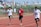 Three young athletes sprint down a red running track, surrounded by a crowd in the background. The runners are dressed in athletic wear, with one wearing a red shirt and the other two in white. The setting appears to be an outdoor sports event on a sunny day.