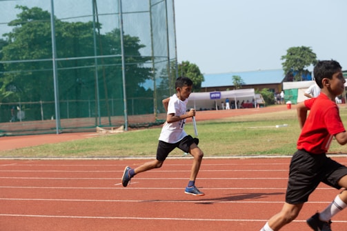 Several children are running on a red track in an outdoor setting. One child in a white shirt and black shorts is in the foreground holding a baton, suggesting a relay race. The background shows green grass, a netted fence, trees, and some buildings with blue rooftops.