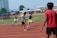 Young athletes are sprinting on a track, engaging in a relay race. The runners are wearing athletic gear, and one of them is holding a baton. The background shows a field and a crowd of spectators in a stadium setup.
