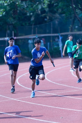 Several young athletes are participating in a relay race on a track. The runners are in motion, with one in the foreground holding a baton and wearing a blue shirt. Other runners are visible further back on the track, dressed in green and blue athletic wear. The setting appears to be outdoors, surrounded by greenery and trees.