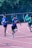 Several young athletes are participating in a relay race on a track. The runners are in motion, with one in the foreground holding a baton and wearing a blue shirt. Other runners are visible further back on the track, dressed in green and blue athletic wear. The setting appears to be outdoors, surrounded by greenery and trees.