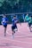 Several young athletes are participating in a relay race on a track. The runners are in motion, with one in the foreground holding a baton and wearing a blue shirt. Other runners are visible further back on the track, dressed in green and blue athletic wear. The setting appears to be outdoors, surrounded by greenery and trees.