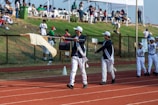 A group of people dressed in coordinated uniforms, including sashes and caps, are marching on a red running track. One person is holding a flag, while others carry signs. Spectators are seated in the background under tents, observing the event from a grassy slope. The atmosphere suggests a formal parade or sporting event.