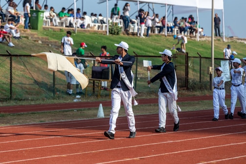 A group of people dressed in coordinated uniforms, including sashes and caps, are marching on a red running track. One person is holding a flag, while others carry signs. Spectators are seated in the background under tents, observing the event from a grassy slope. The atmosphere suggests a formal parade or sporting event.