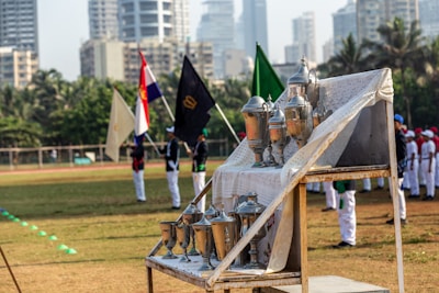 A collection of trophies is displayed on a metal stand covered with a lace cloth. In the background, a group of people in uniforms stands on a field, holding various flags. Tall buildings and palm trees can be seen in the distance.