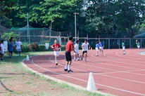 Children are running on a red athletic track surrounded by greenery. The scene is lively with several kids participating in a race, some in the foreground and others in the background. Spectators, likely peers, are standing on the grassy area adjacent to the track. The track is marked with white lines and cones are placed to guide the runners.