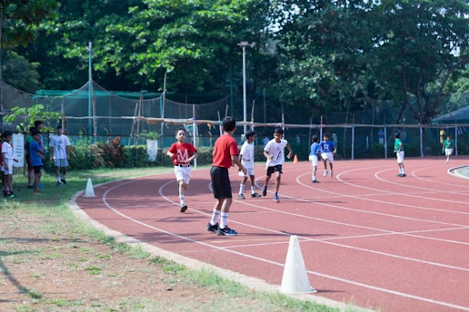 Children are running on a red athletic track surrounded by greenery. The scene is lively with several kids participating in a race, some in the foreground and others in the background. Spectators, likely peers, are standing on the grassy area adjacent to the track. The track is marked with white lines and cones are placed to guide the runners.