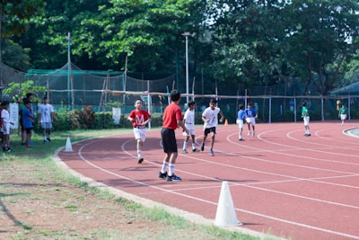 Children are running on a red athletic track surrounded by greenery. The scene is lively with several kids participating in a race, some in the foreground and others in the background. Spectators, likely peers, are standing on the grassy area adjacent to the track. The track is marked with white lines and cones are placed to guide the runners.