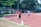 Young boys are participating in a relay race on a red running track. One boy in a red shirt is holding a baton and appears to be leading, while others follow closely behind. Spectators can be seen near the track, all surrounded by trees and greenery.