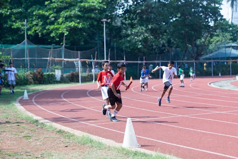Young boys are participating in a relay race on a red running track. One boy in a red shirt is holding a baton and appears to be leading, while others follow closely behind. Spectators can be seen near the track, all surrounded by trees and greenery.