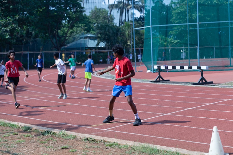 A group of young people are running and walking on a track, wearing numbered T-shirts. The scene is energetic with people in active movement. The environment includes greenery and a building structure in the background.