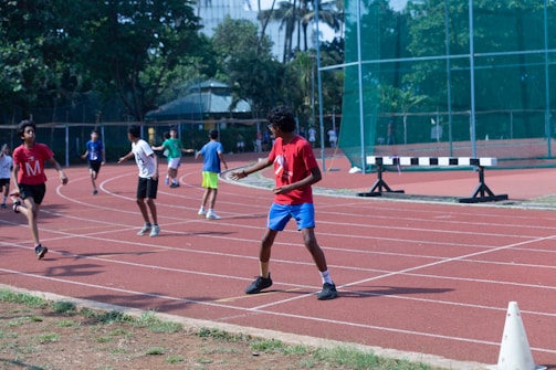 A group of young people are running and walking on a track, wearing numbered T-shirts. The scene is energetic with people in active movement. The environment includes greenery and a building structure in the background.