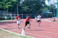 Children are running a relay race on a track field surrounded by greenery and fencing. One child is holding a baton, leading in the race, while others are in pursuit. The scene is set on a clear day, suggesting an outdoor athletic event.