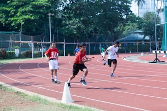 Children are running a relay race on a track field surrounded by greenery and fencing. One child is holding a baton, leading in the race, while others are in pursuit. The scene is set on a clear day, suggesting an outdoor athletic event.