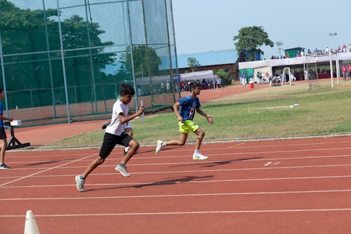 Two athletes are sprinting on a track during a relay race. The runner in the foreground is handing over a baton. A sports field with a cage and spectators in the background is visible under clear skies.