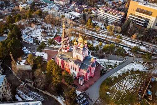 An aerial view of a vibrant, ornately designed Orthodox-style church with red walls and golden domes. The structure is surrounded by trees and pathways, with a scattering of snow on the ground. The surrounding area includes other buildings and a mix of urban and natural landscapes.