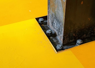 A close-up view of a metal support beam fastened to a vibrant yellow surface. The beam is secured by large bolts and appears weathered, with visible rust and texture. The contrast between the bright, smooth yellow surface and the rough, dark metal creates a striking visual effect.