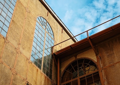 An architectural structure with large arched windows and a textured facade made of metal beams and rivets. The building exhibits an industrial style with a slightly weathered appearance. The sky is clear with a few clouds, adding contrast to the warm tones of the construction.