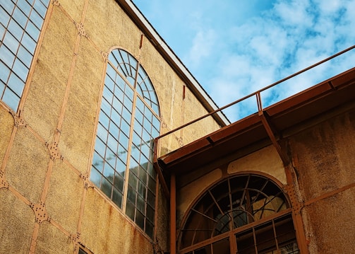 An architectural structure with large arched windows and a textured facade made of metal beams and rivets. The building exhibits an industrial style with a slightly weathered appearance. The sky is clear with a few clouds, adding contrast to the warm tones of the construction.