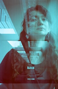 A double exposure photograph featuring a woman with long hair superimposed on an image of a modern architecture stairway with clean lines and geometric patterns. The scene is tinted with a blue hue, giving it a surreal and contemplative feel. The woman is wearing earrings and is looking slightly upwards, adding to the introspective emotional tone.