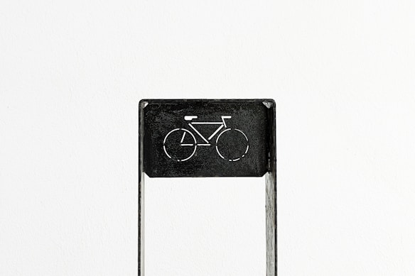 A minimalist black metal sign with a cut-out depiction of a bicycle, set against a plain white background. The design is simple and contrasts sharply with the background.