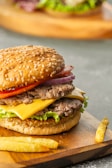 A juicy double cheeseburger with sesame seed bun featuring layers of fresh lettuce, ripe tomato, red onion, and melted cheese, placed on a wooden board with a few french fries. The background shows another blurred burger.