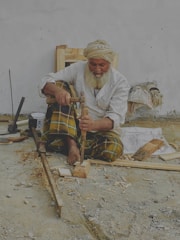 An elderly man with a white beard and turban is sitting on the ground, working with wood tools to carve a piece of wood. He wears a white shirt and plaid pants. Various woodworking tools and wood shavings are scattered around him. He appears to be focused on his craft.