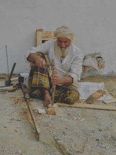 An elderly man with a white beard and turban is sitting on the ground, working with wood tools to carve a piece of wood. He wears a white shirt and plaid pants. Various woodworking tools and wood shavings are scattered around him. He appears to be focused on his craft.