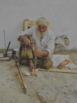 An elderly man with a white beard and turban is sitting on the ground, working with wood tools to carve a piece of wood. He wears a white shirt and plaid pants. Various woodworking tools and wood shavings are scattered around him. He appears to be focused on his craft.