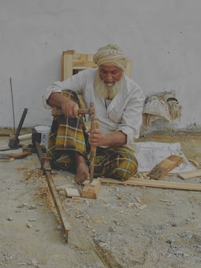 An elderly man with a white beard and turban is sitting on the ground, working with wood tools to carve a piece of wood. He wears a white shirt and plaid pants. Various woodworking tools and wood shavings are scattered around him. He appears to be focused on his craft.