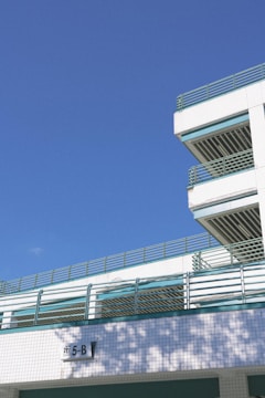 An outdoor structure, likely a multi-story parking garage, with green metal railings and white tiled walls. Shadows of trees are visible on the building facade. The sky is clear and blue, and a sign with the notation '5-B' is visible.
