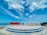 A large sign with the words 'TIANG BENDERA' stands on a tiered circular platform by the seaside. The sky is bright and partly cloudy, and the ocean can be seen stretching out in the background.
