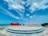 A large sign with the words 'TIANG BENDERA' stands on a tiered circular platform by the seaside. The sky is bright and partly cloudy, and the ocean can be seen stretching out in the background.