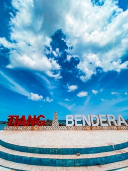 A landscape with a prominently featured structure displaying large letters spelling 'TIANG BENDERA'. The sky above is expansive and bright blue, filled with fluffy white clouds. The foreground has a circular tiled pattern, leading towards the structure.