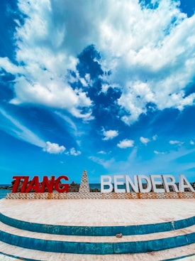 A landscape with a prominently featured structure displaying large letters spelling 'TIANG BENDERA'. The sky above is expansive and bright blue, filled with fluffy white clouds. The foreground has a circular tiled pattern, leading towards the structure.