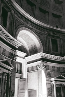 A black and white photograph capturing the architecture of a grand interior space with a large arched doorway, intricate columns, and detailed molding. The light streams through the doorway, highlighting the detailed carvings and architectural elements.