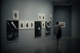 A person is walking through a dimly lit art gallery. The walls display a series of monochrome photographic artworks, arranged in a neat layout. Some images appear to depict telescopes and individuals in various poses.