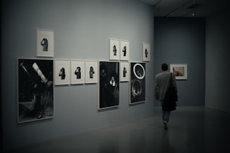 A person is walking through a dimly lit art gallery. The walls display a series of monochrome photographic artworks, arranged in a neat layout. Some images appear to depict telescopes and individuals in various poses.