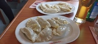 Two plates of boiled dumplings are placed on a wooden table. The dumplings appear to be filled and are accompanied by drinks, including a glass of beer.