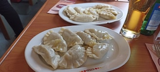 Two plates of boiled dumplings are placed on a wooden table. The dumplings appear to be filled and are accompanied by drinks, including a glass of beer.