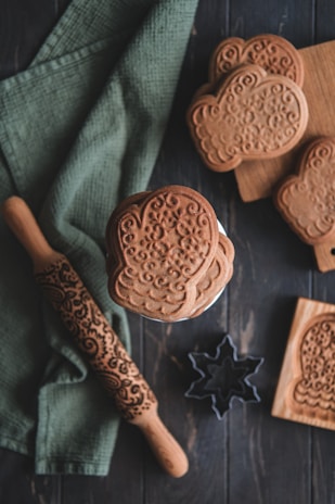 A collection of intricately patterned cookies is displayed alongside a decorative rolling pin and wooden molds. The cookies have a textured, floral design, and are placed on a rustic wooden surface. A green cloth is draped beneath the items, adding a touch of color. A star-shaped cookie cutter and wooden cutting board complete the cozy baking scene.