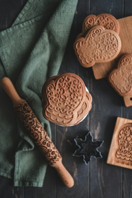 A collection of intricately patterned cookies is displayed alongside a decorative rolling pin and wooden molds. The cookies have a textured, floral design, and are placed on a rustic wooden surface. A green cloth is draped beneath the items, adding a touch of color. A star-shaped cookie cutter and wooden cutting board complete the cozy baking scene.
