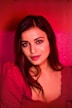 A woman with long dark hair is wearing a red top. The background is a vibrant red with pink hues, contributing to a warm and inviting atmosphere. Her expression is soft and slightly smiling, with well-defined eyebrows and a subtle makeup look.