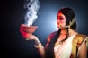 A person dressed in traditional attire holds a clay incense burner with smoke rising from it. The individual wears ornate jewelry, including a nose ring and headpiece, and is adorned in a white and gold sari. The lighting creates a dramatic contrast with a dark background, highlighting the colorful makeup and intricate details of the garments.